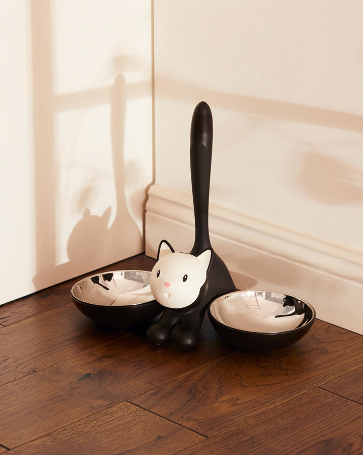Der doppelte Katzennapf Tigrito für Futter und Wasser zeigt eine schwarz-weiße Katze, die von zwei Schalen flankiert wird, die außen aus schwarzem Thermoplast und innen aus hygienischem Edelstahl bestehen. Ideale Alternative zu langweiligen Futternäpfen für Katzenbesitzer